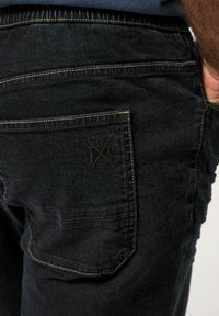 Jeans de mezclilla negra con acabado texturizado, que cuentan con un bolsillo trasero con logo en relieve y costuras amarillas contrastantes en los bordes.