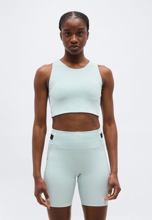 Mujer con top corto sin mangas de color azul claro y pantalones ciclistas de talle alto a juego, sobre un fondo blanco liso.