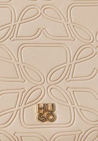 Beige kunstlær med preget sløyfedesign. Gull "HUGO"-logo festet, som gir en elegant aksent. Glatt tekstur, moderne utseende.