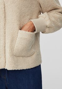Beżowa polarowa kurtka o textured powierzchni, z żeberkowym mankietem i boczną kieszenią. Luźny krój i miękki materiał.