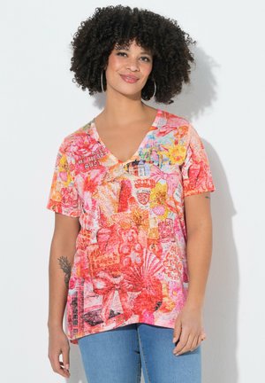 Kobieta o kręconych włosach, nosząca duże kolczyki-koła oraz kolorową bluzkę z krótkim rękawem i dekoltem w serek, zdobioną kwiatowymi i graficznymi wzorami.