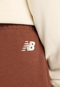 Bruine trainingsbroek met een wit 'New Balance'-logo op de dij, gemaakt van zacht materiaal met een geribde tailleband. Klassiek, eenvoudig ontwerp.
