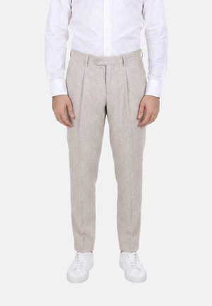 Uomo che indossa pantaloni beige plissettati, camicia bianca abbottonata e sneakers bianche, in piedi davanti a uno sfondo semplice.