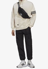 Svart Adidas midjeväska som bärs diagonalt över en beige hoodie med tre svarta ränder, kombinerat med svarta cargobyxor och vita sneakers.