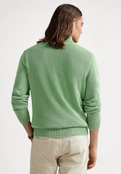 Zöld kötött pulóver bordázott gallérral és mandzsettákkal. Texturált anyaggal és laza szabással rendelkezik. Világos bézs nadrággal viselve.