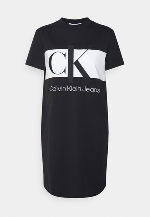Czarna sukienka z krótkim rękawem typu t-shirt z białym panelem na wysokości klatki piersiowej, z dużymi literami "CK" i tekstem "Calvin Klein Jeans" poniżej.