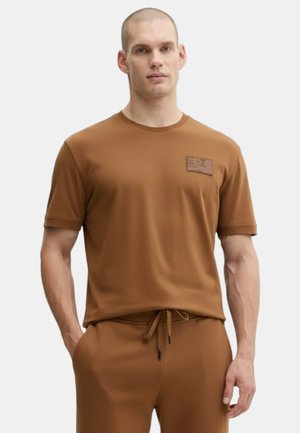 Uomo che indossa una camicia marrone a maniche corte con toppa sul petto e pantaloni con coulisse abbinati, in piedi con una mano in tasca.