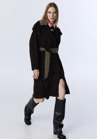 Vrouw in een donkeren bruine jas met riem en geruite accenten, zwarte kniehoge laarzen en een roze blouse met kraag, staand tegen een effen achtergrond.