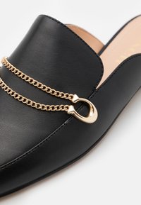 Svart lädersko med slip-on design som har en guldkedjeaccent och en dekorativ detalj i metall. Skon har en slät yta och en rund form.