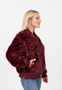 Giacca bomber in pelliccia sintetica bordeaux con polsini e orlo a coste, tasche laterali e una zip dorata. Presenta una finitura morbida e texture.