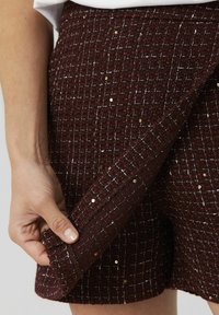 Jupe en tweed bordeaux avec un motif texturé, comprenant des fils blancs et des sequins. Une main tire sur l'ourlet pour montrer son matériau et son design.