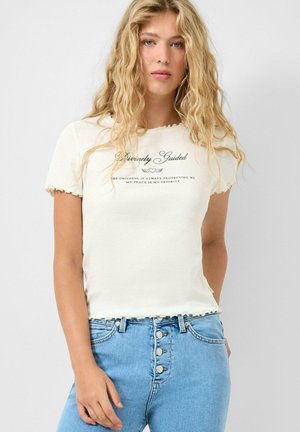 Junge Frau mit langen, blonden, welligen Haaren trägt ein cremefarbenes T-Shirt mit Schriftzug und hellblaue, hoch taillierte Jeans mit sichtbaren Knöpfen.