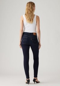 Kvinne med langt blondt hår, iført en hvit tanktopp, mørkeblå skinny jeans og svarte høye sandaler, som står med ryggen til mot en ensfarget bakgrunn.