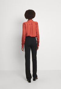 Kvinna med afrohår som bär en röd långärmad blus, svarta högmidjade byxor och svarta klackskor, stående framför en vit bakgrund.