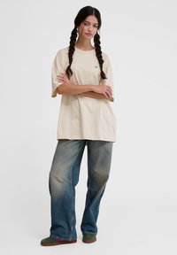 Beige oversized t-shirt med korte ærmer og grøn broderi, parret med lyseblå baggy jeans og grønne og brune sko.