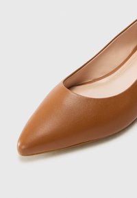 Chaussure en cuir marron à bout pointu, avec une texture lisse et un design court, révélant une doublure intérieure claire et une forme minimaliste.