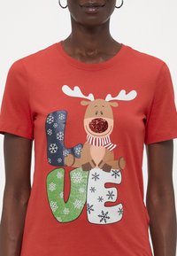 Moteris dėvi raudoną marškinį su šventiniais elniukais ir įvairiaspalvėmis "MEILĖ" raidėmis, dekoruotomis snaigėmis ir raudonu blizgiu nosimi.