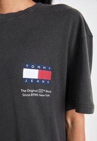 Camiseta de algodón gris con un logo rectangular de color que presenta el texto "TOMMY JEANS" y pequeños detalles de la marca impresos debajo. Mangas cortas.