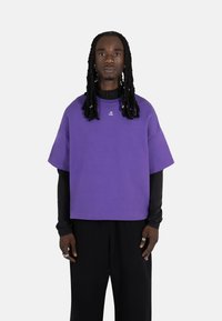 Lilla oversized t-skjorte med korte ermer, laget av myk stoff. Har en liten logo på brystet og blir brukt over en svart langermet skjorte.