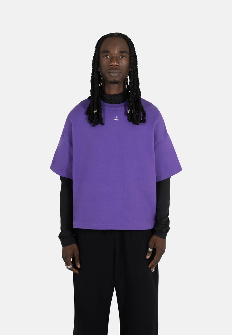 Lilla oversized t-skjorte med korte ermer, laget av myk stoff. Har en liten logo på brystet og blir brukt over en svart langermet skjorte.
