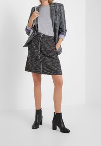 Harmaa ruudullinen bleiseri harmaan t-paidan päällä, yhdistettynä teksturoituun mustaan hameeseen, jossa on hopea vetoketju, ja mustiin nilkkasaappaisiin.