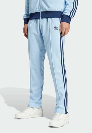 Mężczyzna ubrany w jasnoniebieskie spodnie dresowe Adidas z ciemnoniebieskimi paskami i białe buty sportowe, stojący na jednolitym tle.