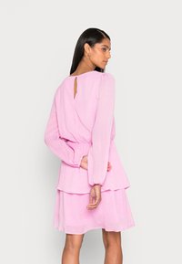 Vestido rosa, em camadas, com mangas longas, elástico na cintura e um recorte em forma de gota nas costas. O material apresenta um padrão de pontos subtil.