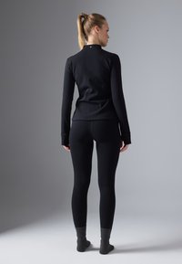 Melna garām piedurknēm topiņš ar augstu apkakli un pieguļošu griezumu, kombinēts ar melnām leggingām un tumši pelēkām zeķēm. Gluda tekstūra, minimāls dizains.