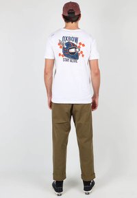 T-shirt blanc avec un graphisme de casque coloré et le texte "STAY ALIVE" au dos, associé à un pantalon vert olive et des baskets noires.