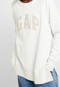 Sudadera blanca con un logo "GAP" en relieve en color beige, mangas raglán y aberturas laterales en el dobladillo. Fabricada con material suave y con un corte relajado.