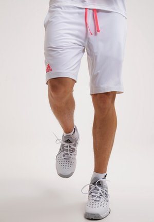 Mężczyzna w białych spodenkach Adidas z różowym sznurkiem i białych butach Adidas, idący z uniesioną jedną stopą w trakcie kroku.