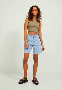 Lichtblauwe denim shorts met een raw hem, gecombineerd met een olijfgroene tanktop. Model draagt zwarte sandalen en ronde oranje zonnebril.