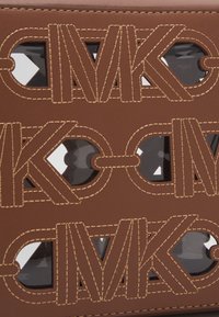 Brun lædertaske med udskåret logodesign og kontrasterende syninger. Dekorativ hardware synlig gennem udskæringerne. Skinnende indvendigt materiale.