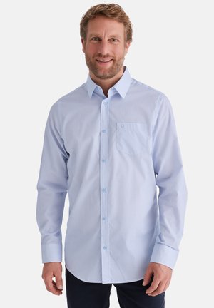 Lächelnder Mann mit kurzen braunen Haaren und Bart trägt ein hellblaues Hemd mit Brusttasche und steht vor einem schlichten weißen Hintergrund.