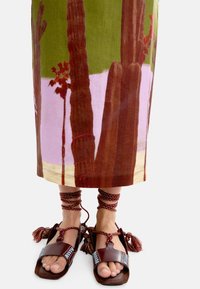 Brązowe skórzane sandały z kolorowymi frędzlami i dekoracyjnymi sznurówkami, w połączeniu z długą spódnicą z printem kaktusa i krajobrazu w odcieniach zieleni i różu.