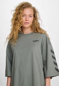 Olijfgroene oversized t-shirt met een ronde hals, korte mouwen en een zwart logo op de borst. Heeft zwarte chevrons op de mouwen.