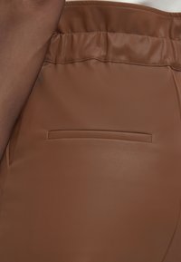 Κοντινή λήψη καφέ παντελονιών από συνθετικό δέρμα με ελαστική μέση και οριζόντια σχισμή τσέπης στο πίσω μέρος.