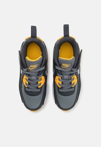 Nike Air Max teniske z sivo zgornjo plastjo, rumenimi poudarki, sivimi vezalkami, oblazinjenim jezikom in gumijastim podplatom. Imajo teksturirano površino in detajl z logotipom.