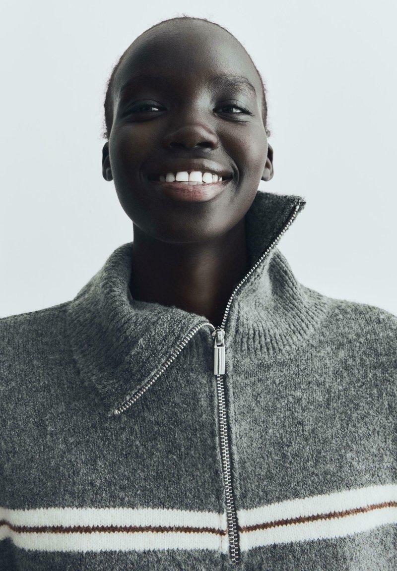 Sivi dukserica sa zatvaračem i visokim ovratnikom; sadrži bijele i smeđe pruge preko prsnog dijela; izrađena od teksturiranog pletenog materijala.