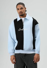 Pull-over surdimensionné à design bicolore noir et bleu clair, avec col zippé et détail brodé "Saintessence". Textile en mélange de coton.