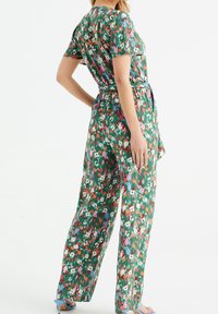 Květinový overal s krátkými rukávy, zelený podklad, živé květy v červené, růžové a modré barvě, s detaily na převázání v pase. Nosený s modrými sandály.