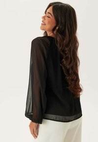 Blusa nera trasparente con maniche lunghe e fluide. Tessuto leggero, texture liscia e vestibilità ampia, con un orlo arrotondato sul retro.
