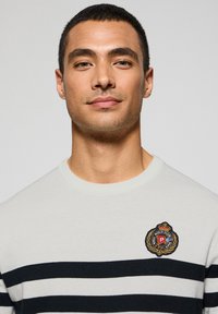 Ljusgrå tröja med marinblå horisontella ränder, rund halsringning och broderad logotyp på vänster bröst. Mjuk stickad textur.