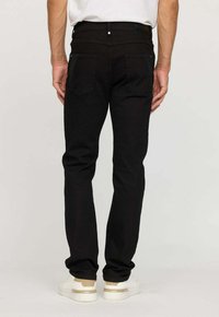 Homme portant un jean slim noir et des baskets blanches, se tenant les mains détendues sur les côtés devant un fond uni.