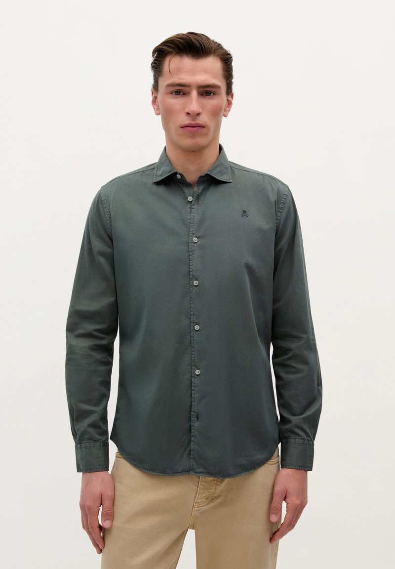 Camisa de manga larga verde oscura con cuello abotonado, logo sutil y una textura suave. Corte ajustado con dobladillo curvado y ligeros detalles de botones.