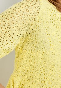 Top de encaje en amarillo claro, con patrones florales, mangas cortas y tejido texturado. Acabado suave con detalles sutiles.