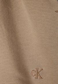 Beżowa teksturowana tkanina z wzorem waflowym, z wyszywanym ciemnobeżowym logo "CK" w dolnym prawym rogu.