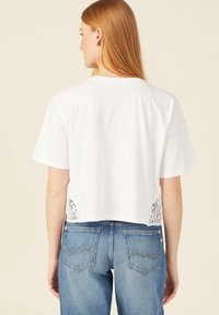 Witte cropped t-shirt met korte mouwen, verfraaid met kantdetails aan de zijkanten. Gecombineerd met blauwe jeans, wat een casual uitstraling geeft.