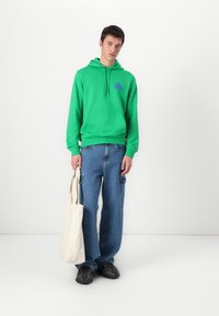 Felpa verde con cappuccio e tasca frontale, abbinata a jeans cargo blu a vestibilità ampia e una borsa a spalla color crema. Sneakers nere indossate.