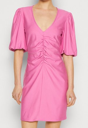 Nainen pukeutuneena vaaleanpunaiseen mekkoon, jossa on pöyhistetyt lyhyet hihat ja etuosassa rypytys, seisoo yksiväristä taustaa vasten.